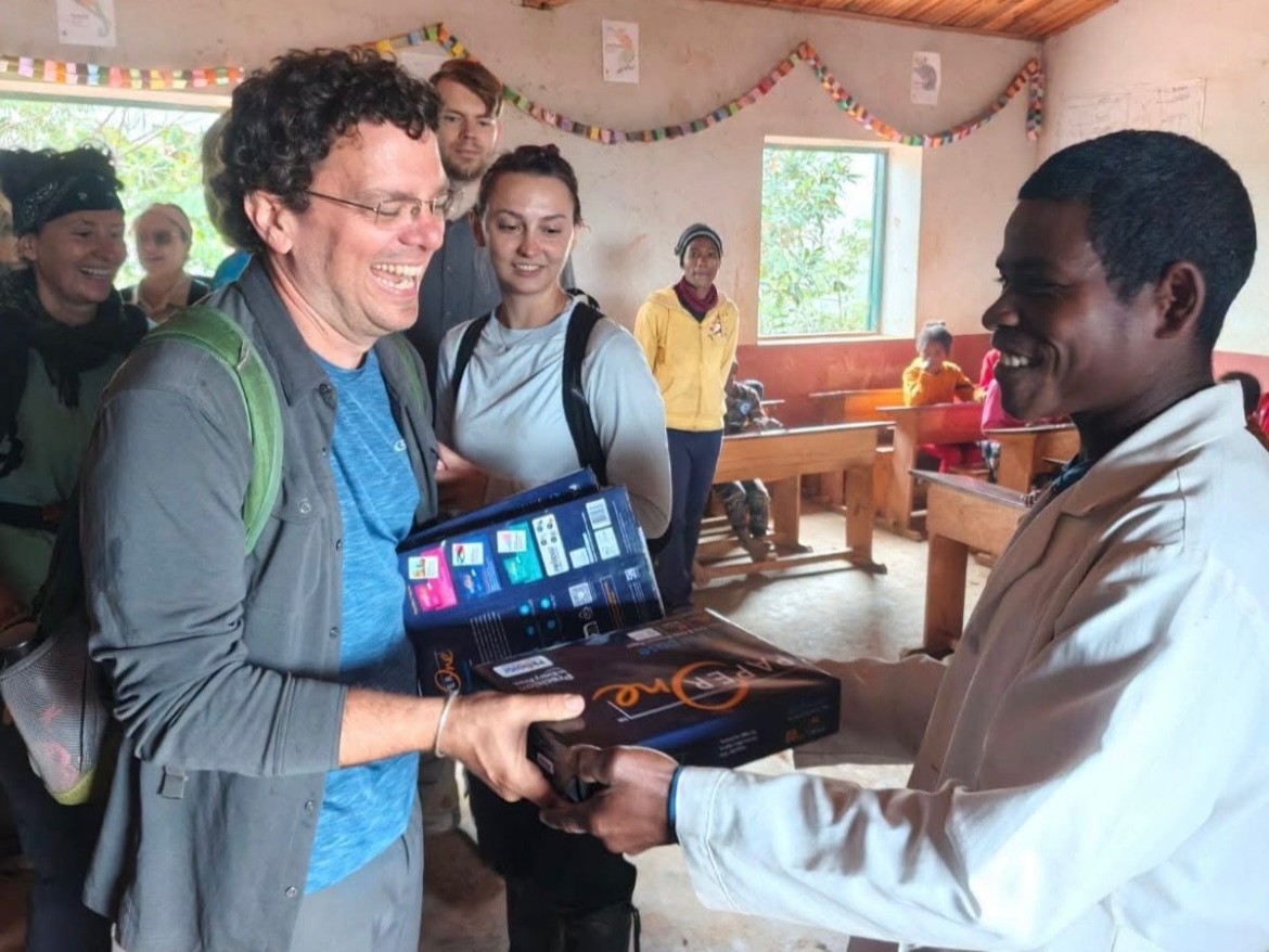 Man smiles while handing over supplies to another man in a classroom with others observing.