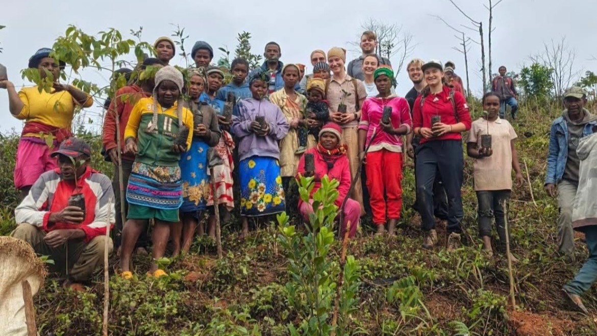 Group of people standing outdoors on a hillside, holding plants and tools, surrounded by greenery.