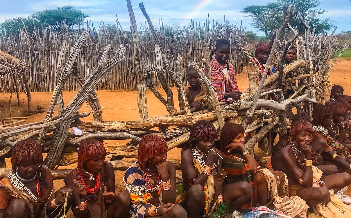Group of people sitting near a wooden fence in a rural area under a cloudy sky with a faint rainbow.